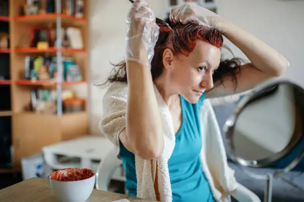 Woman is coloring her own hair at home. During lockdown period she is staying at home and can't go to hair salons. She wears hand gloves and has small container with hair color and a mirror in front of her. She is holding hair coloring brush and is placing color on her roots.