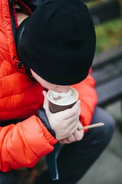 November 21, 2020 - Warsaw, Poland: teenage boy bending, sipping milky coffee from a paper cup while sitting on a bench, wearing a red jacket and black hat, face obscured.