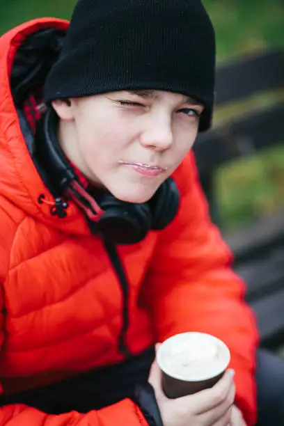 November 21, 2020 - Warsaw, Poland: face of a teenage boy in red jacket, black hat and headphones holding cappuccino in his hand, looking at camera, smiling naughty, winking.