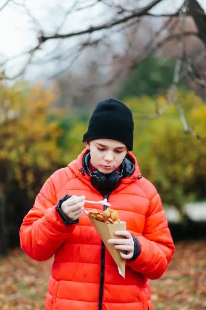 November 21, 2020 - Warsaw, Poland: Serious concentrated teen boy in red jacket and hat is standing, eating his bubble waffle (hong kong wafer) with whipped cream looking a bit sad, lonely in a park)