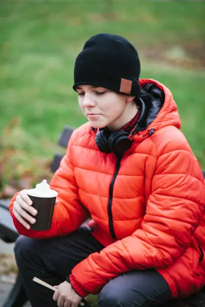 November 21, 2020 - Warsaw, Poland: cute teenager boy preparing to sip and taste his coffee in a cup with whipped cream on top while sitting in a park in warm clothes.