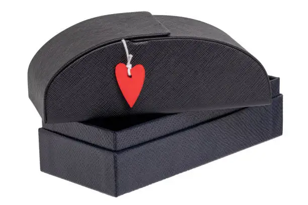 Closeup of a closed sturdy (hardcase) black luxury leather glasses case with a small red heart on an opened gift box isolated on white. Valentine's Day concept. Macro.
