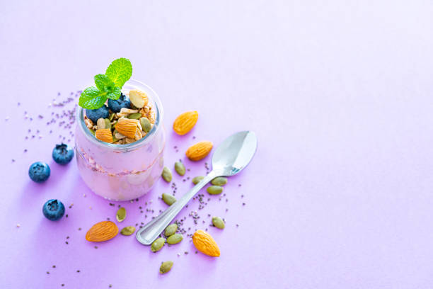 Healthy food backgrounds: high angle view of two yogurt glass containers with blueberries, almonds, pumpkin seeds and chia seeds shot on pastel purple background. Goji berries, blueberries, almonds, pumpkin seeds and chia seeds are around the yogurt containers. The composition is at the right of an horizontal frame leaving useful copy space for text and/or logo at the left. High resolution 42Mp studio digital capture taken with Sony A7rII and Sony FE 90mm f2.8 macro G OSS lens