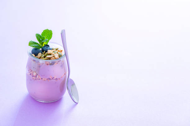 Healthy food backgrounds: yogurt glass containers with blueberries, almonds, pumpkin seeds and chia seeds shot on pastel purple background. A metal spoons is beside the yogurt container. The composition is at the left of an horizontal frame leaving useful copy space for text and/or logo at the right. High resolution 42Mp studio digital capture taken with Sony A7rII and Sony FE 90mm f2.8 macro G OSS lens