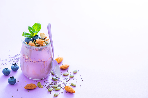 Healthy food backgrounds: high angle view of a yogurt glass container with blueberries, almonds, pumpkin seeds and chia seeds shot on pastel purple background. Blueberries, almonds, pumpkin seeds and chia seeds are around the yogurt container. The composition is at the left of an horizontal frame leaving useful copy space for text and/or logo at the right. High resolution 42Mp studio digital capture taken with Sony A7rII and Sony FE 90mm f2.8 macro G OSS lens