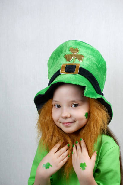 istock Saint Patricks Day. Endearing smiling small girl with decorative red beard, green shamrok leaf on her cheek and leprechaun hat, raised her hands with green nails and clover leaves to beard 1299932685