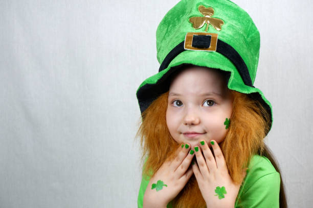 istock Saint Patricks Day celebration. Nice pensive little girl with decorative red beard, green shamrok leaf on her cheek and leprechaun hat, raised her hands with green nails and clover leaves to her face 1299932992