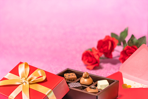 Valentine or white day greeting card depicting a chocolate box adorned with a golden ribbon and a love letter with red roses flowers on a glittering pink background.