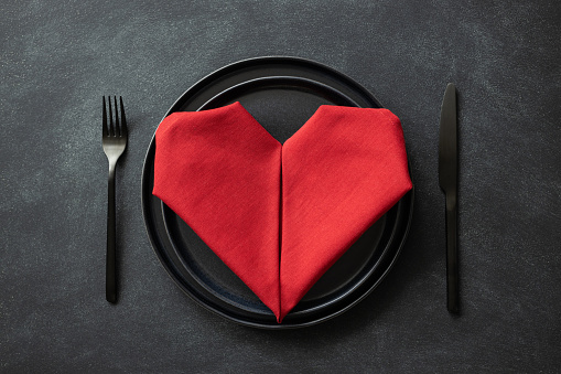 Heart made from red napkin on black plates with black cutlery on black table. Top view on Valentine's day table setting.