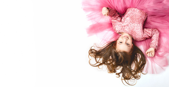little Princess. Portrait of a cute Caucasian little laughing girl in an evening bright pink dress lies on a white background. Horizontally. top view