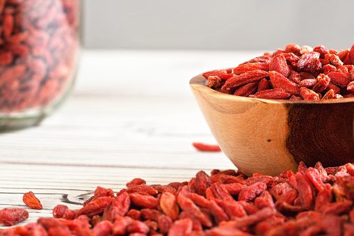 Dried goji berries in wooden bowl, scattered over white boards table under, blurred large glass jar full of fruits in background