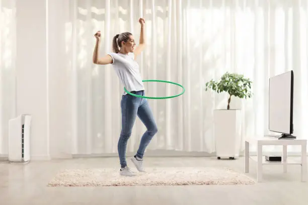 Young woman spinning hula hoop at home in a living room in front of tv