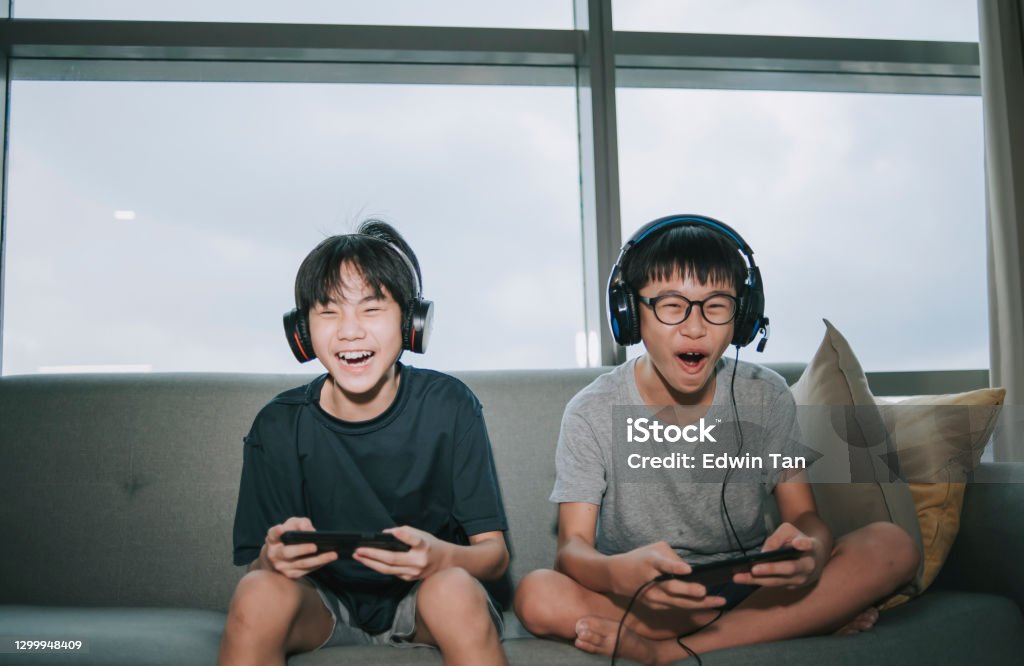 2 asian chinese sibling brother sitting on sofa playing online video games with headset in living room during weekend Video Game Stock Photo