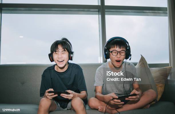 2 Asian Chinese Sibling Brother Sitting On Sofa Playing Online Video Games With Headset In Living Room During Weekend Stock Photo - Download Image Now