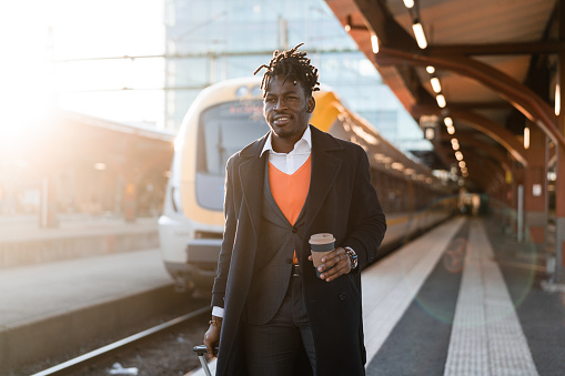 Handsome confident businessman on his way home from work. He is walking on the train station platform at sunset, with a cup of coffee in his hand.