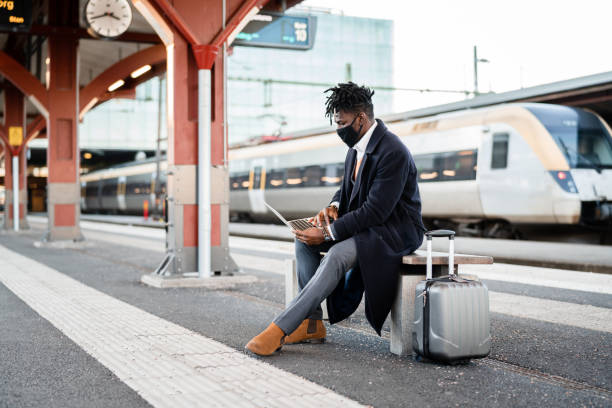 Businessman with face mask working at the train station Handsome confident businessman on his way home from work. He is standing on the train station platform at sunset, waiting for his train. railroad station platform stock pictures, royalty-free photos & images