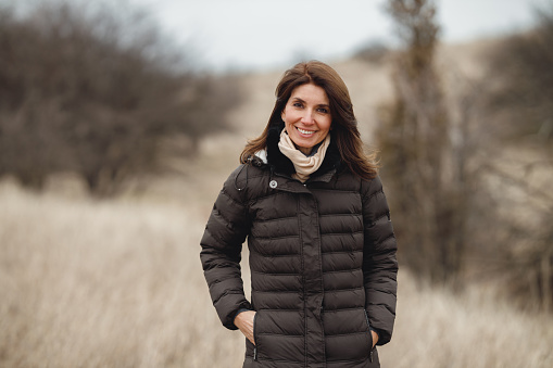 A beautiful mature woman is smiling and posing in a field of dried grass on a cold, windy day.