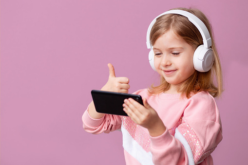 Little girl blogger influencer records videos from the blog on a smartphone, communicates with subscribers, puts likes, listens to music with headphones, remote learning on an isolated background. High quality photo