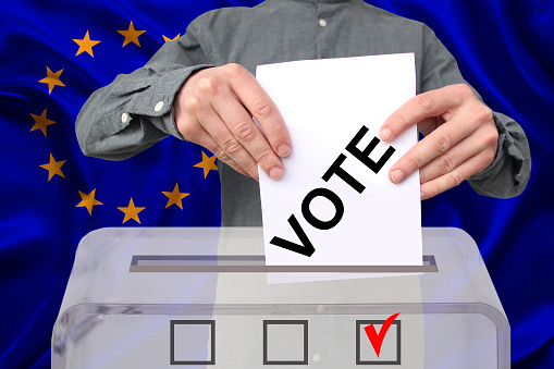 male voter drops a ballot in a transparent ballot box against the background of the European Union national flag, concept of state elections, referendum