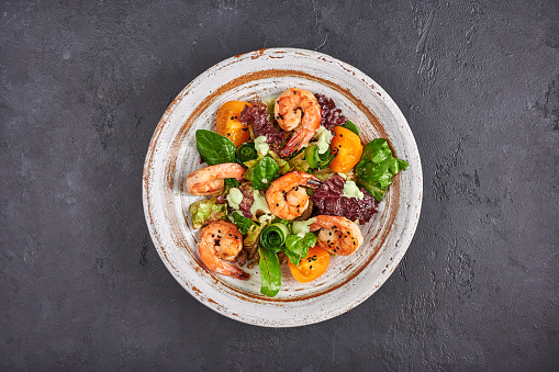 Homemade salad with fried shrimp, yellow tomatoes, arugula, spinach, lettuce and sauce on a ceramic bowl on dark background, top view, copy space