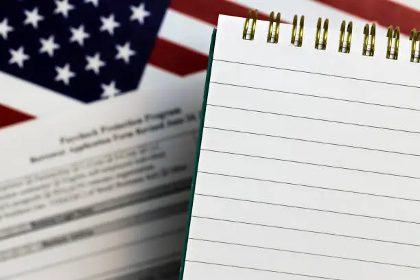 Photo of selective focus photo of open notepad with blank space, on a background of paycheck protection program borrower application form and the United States flag. paycheck protection program new round.