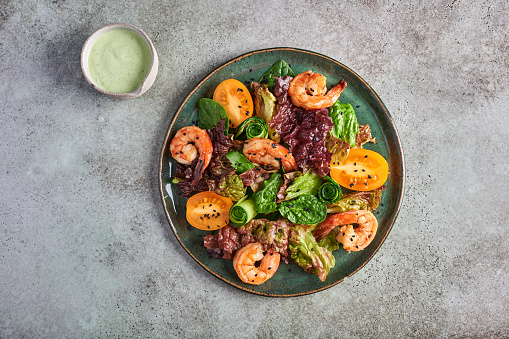 Homemade salad with fried shrimp, yellow tomatoes, arugula, spinach, lettuce and sauce on a ceramic plate on texture background, top view, copy space