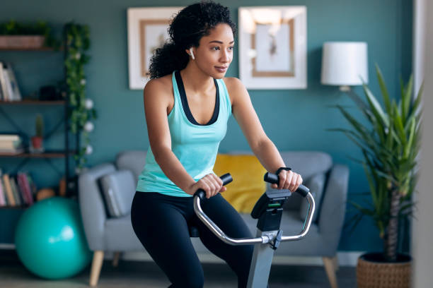 mujer joven africana deportiva haciendo ejercicio en bicicleta estática inteligente y escuchando música en casa. - bicicleta estática fotografías e imágenes de stock