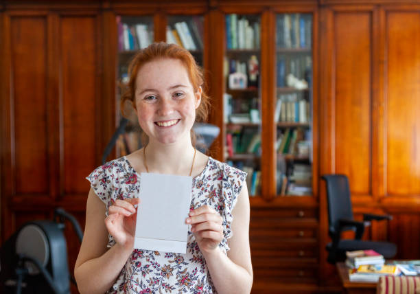 Teenage girl looking at the camera showing a birthday card and is opening birthday presents for her 16th birthday in the living room at home and unwrapping gift boxes stock photo