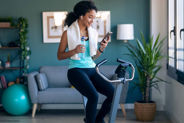 afro joven chica de fitness usando el teléfono móvil mientras entrena en bicicleta de ejercicio en casa. - bicicleta estática fotografías e imágenes de stock