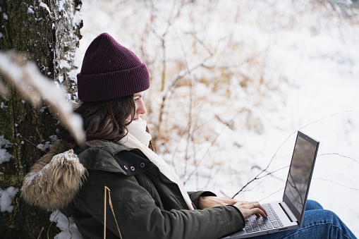 Young girl, woman working in laptop. Walking in beautiful winter forest among trees, firs, covered with snow. Magnificent nature and views. Fashionable image, clothes, parka, hat, mittens, blue jeans.