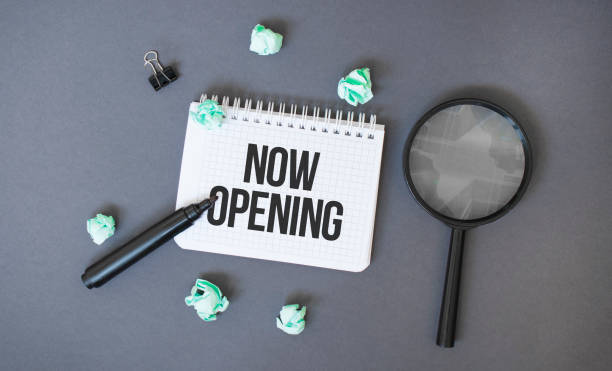 istock Paper notepad with text NOW OPENING, crumpled papers sheets and magnifier glass on dark background 1299961576