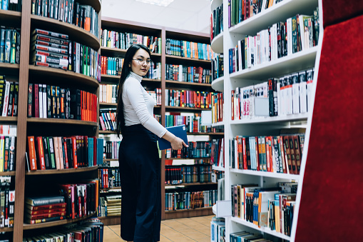 Side view of attractive young woman in glasses with long black hair standing with book in hand surrounded by bookshelves in library and looking at camera
