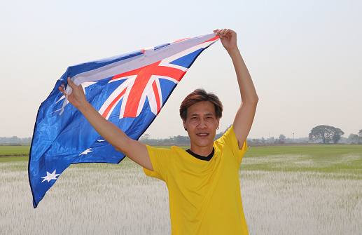 The man in yellow shirt is holding Australia flag in his hands and raising to the end of the arm at the back on nature view and rice fields background.