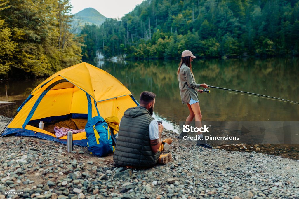 We always make time for fishing Young woman out fishing by a river while his boyfriend making company Camping Stock Photo