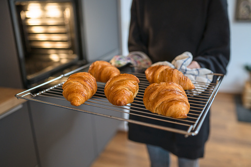 Unrecognisable person holding baking tray and taking baked croissants out of oven.