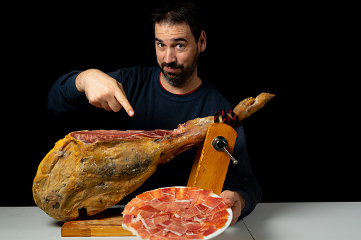 Bearded man with a ham knife ready to cut a piece of typical Spanish ham isolated on black studio background. Super foods. Delicious appetizer. Feeding concept