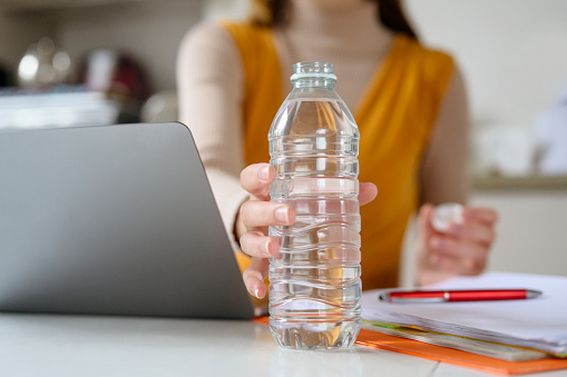 Surface level close-up of Caucasian female telecommuter reaching for uncapped bottle of water as she takes a break from using laptop.