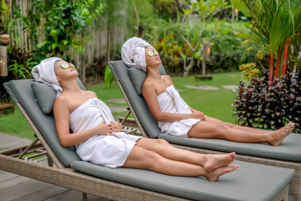 Two Asian friends relaxing on weekend spa getaways Side view shot of two Asian women wrapped in towels and lying on the sunbeds with refreshing cucumber slices on their eyes. They're relaxing on their outdoor villa, enjoying a wellness spa weekend trip. vacation rental mask stock pictures, royalty-free photos & images