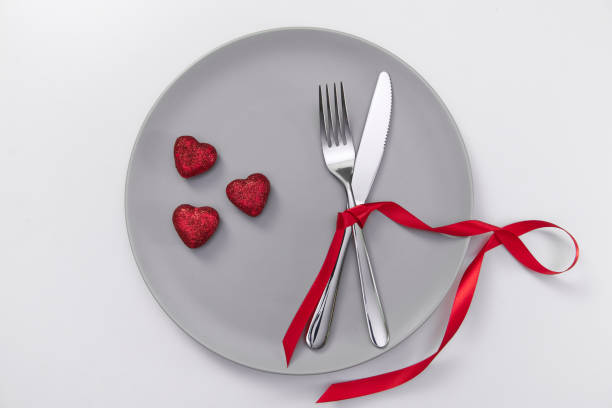 istock Valentine's day festive table setting, flat lay with red heart shape on gray plate, fork, knife and red ribbons on white table. Love dating concept. Place your text and copy space 1299985056