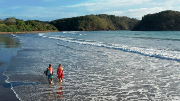 istock Mature couple walk down beach before morning surf session 1299986848