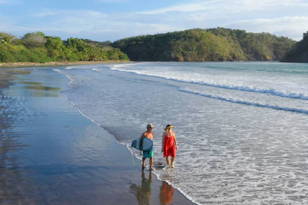 istock Mature couple walk down beach before morning surf session 1299986991