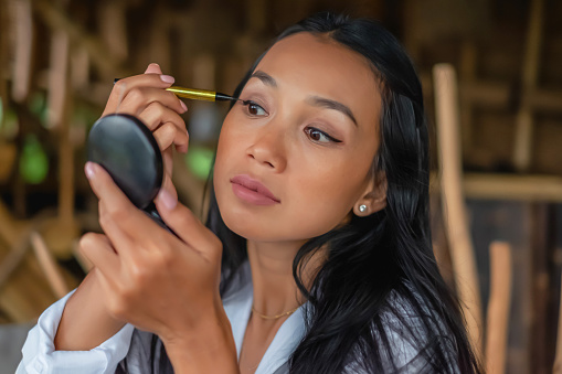 Close up shot of an attractive Indonesian woman looking to her compact pocket mirror and applying black eyeliner into her eyelid during staycation in  bamboo holiday villa.