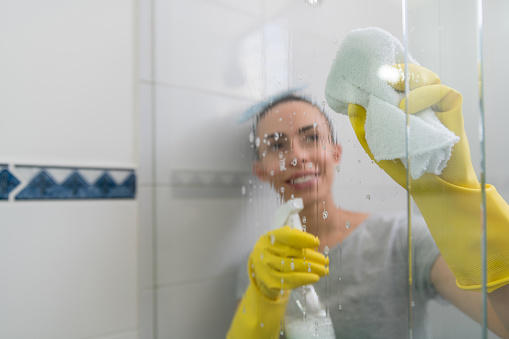 Latin American woman at home in the shower cleaning the glass using cleaning products and a cloth - domestic life concepts