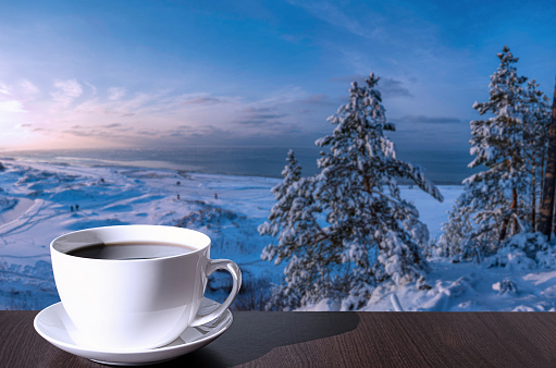 Cup of coffee on the table with view of snowy beach with dunes, Baltic sea and covered in snow fir trees on the hill.