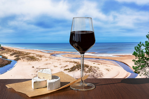 Glass of red wine with brie cheese against view of beach and Baltic see. Beach with white sand, pine tree and blue sea or ocean.