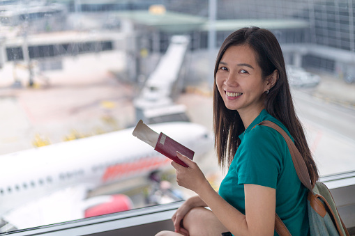 Mid aged woman waiting for her flight at the airport lounge, holding a passport and boarding pass in hand and smiling at the camera.