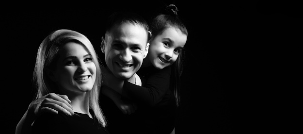 Joyful family mother, father and little girl in black clothes with dark background. Family portrait.