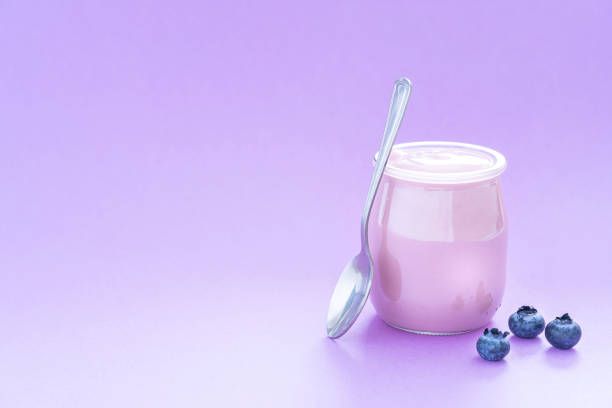 Healthy food backgrounds: front view of a yogurt glass container shot on purple background. A metal spoon is beside the yogurt container. The composition is at the right of an horizontal frame leaving useful copy space for text and/or logo at the left. High resolution 42Mp studio digital capture taken with Sony A7rII and Sony FE 90mm f2.8 macro G OSS lens