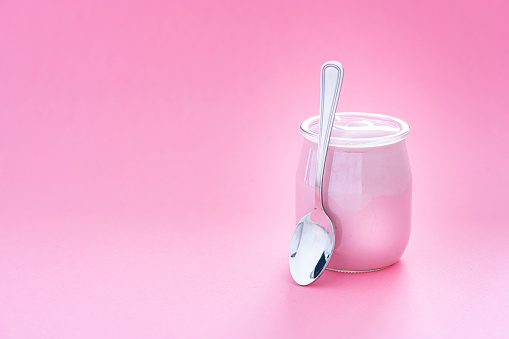 Healthy food backgrounds: front view of a yogurt glass container shot on pastel pink background. A metal spoon is beside the yogurt container. The composition is at the right of an horizontal frame leaving useful copy space for text and/or logo at the left. High resolution 42Mp studio digital capture taken with Sony A7rII and Sony FE 90mm f2.8 macro G OSS lens