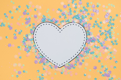 Paper elements in shape of white heart with copy space on orange background surrounded by small colorful flowers. Symbols of love for Happy Women's, Mother's, Valentine's Day